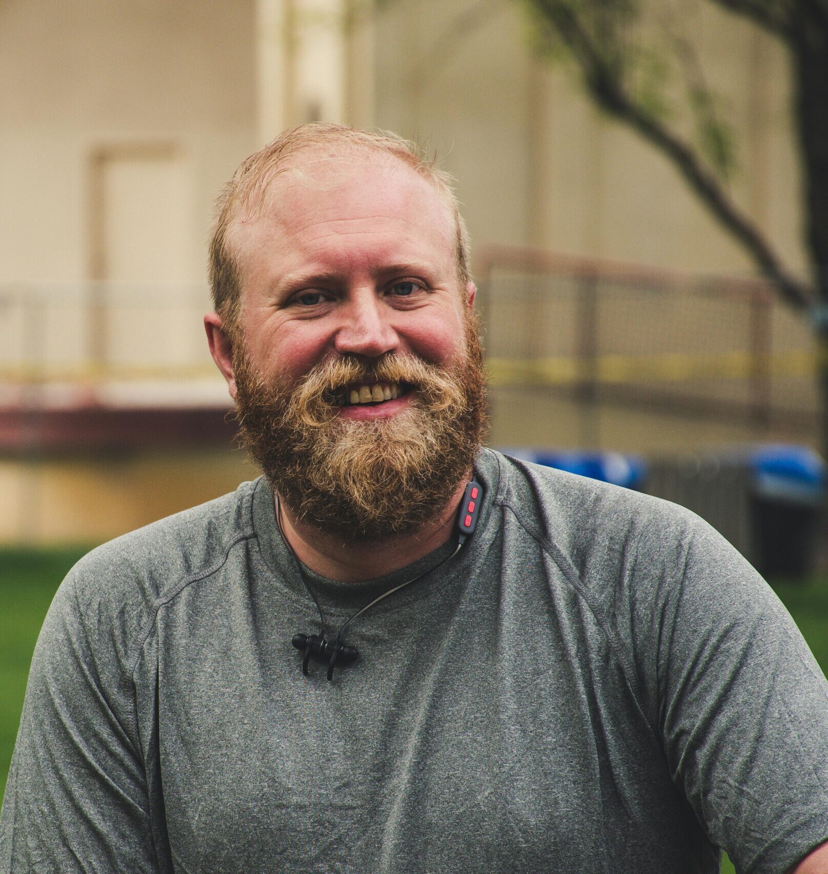 A cheerful bearded man smiling outdoors in a casual setting with greenery.